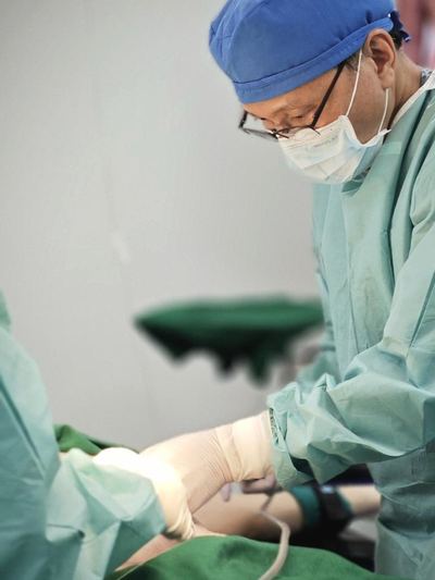 Operative photograph of a surgeon in blue cap and gown working at a draped field. The patient's face is not shown.
